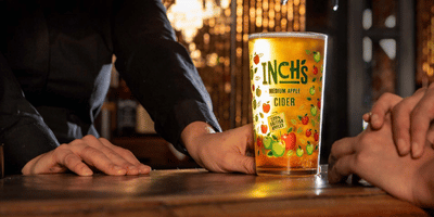 A pint of Inch’s Apple Cider on a bar counter, held at the base by a person’s hand. The glass is filled with golden cider and decorated with colourful apple illustrations and the Inch’s branding. Warm, glowing lights blur in the background A pint of Inch’s Apple Cider on a bar counter, held at the base by a person’s hand. The glass is filled with golden cider and decorated with colourful apple illustrations and the Inch’s branding. Warm, glowing lights blur in the background