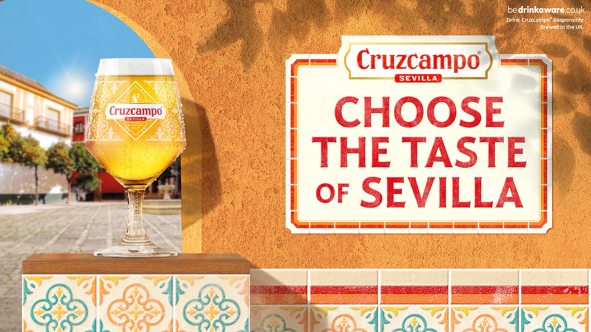 Cruzcampo beer showing a glass of golden beer beside a sign reading Choose the Taste of Sevilla with a sunlit Spanish-style street and decorative tiles in the background. Cruzcampo beer showing a glass of golden beer beside a sign reading Choose the Taste of Sevilla with a sunlit Spanish-style street and decorative tiles in the background.